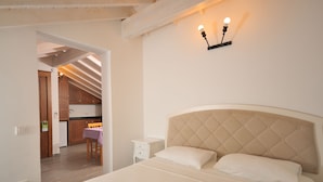 A bedroom with a large bed, a nightstand, a small table with a chair, and a kitchenette in the background. - Brothers Cesme Hotel (Cesme)