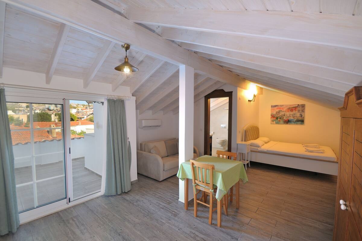 A cozy attic bedroom with a bed, a small table with a chair, a sofa, and a view through a sliding door.