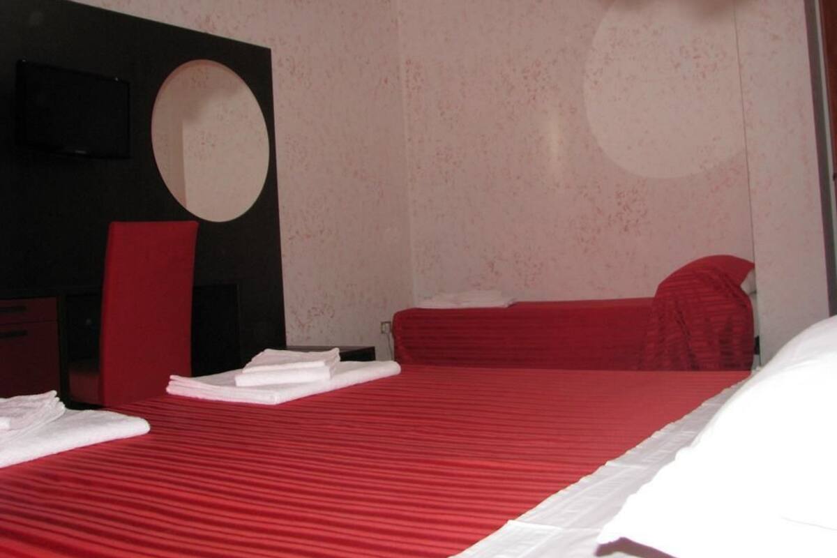 A room with a red bed, white pillows, and a red chair. There is a round mirror on the wall and a TV mounted on the wall.