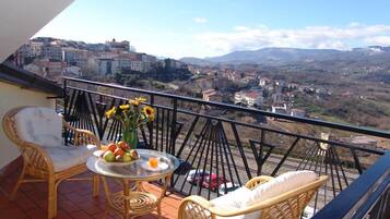 Ein Balkon mit Korbmöbeln, einem Tisch mit Früchten und einer Vase, sowie Blick auf eine Stadt und Berge.