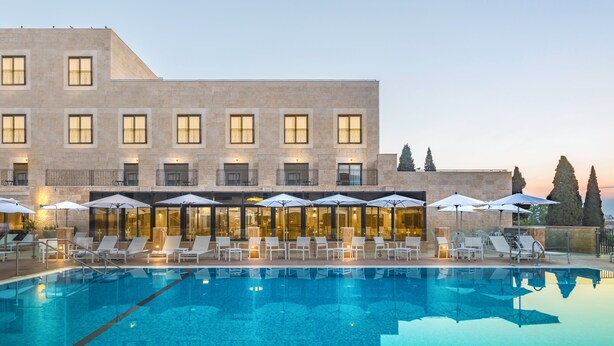 A swimming pool with lounge chairs and umbrellas, a building with multiple windows and balconies, and a clear sky. - Edmond Luxury Hotel & Spa (Rosh Pinna)
