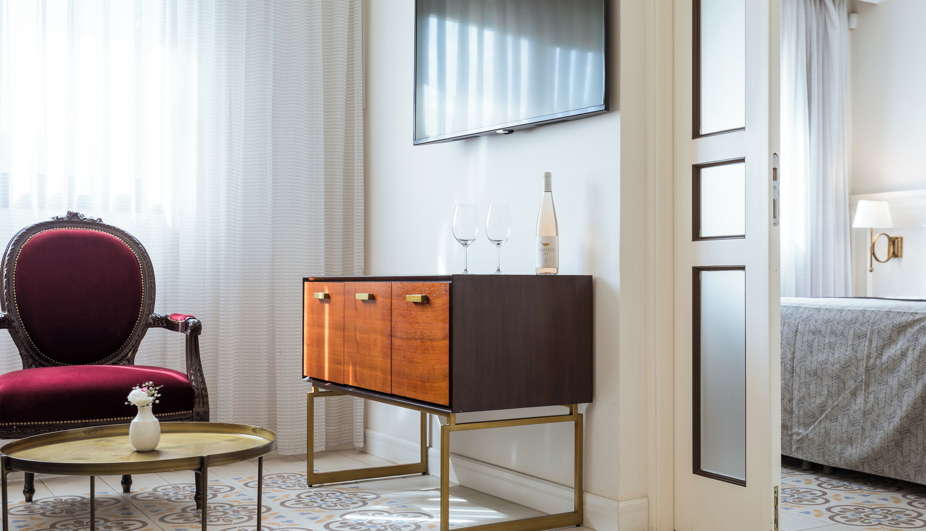 Habitación de hotel con un sillón rojo, un mueble de madera con dos cajones, una mesita redonda con un florero, un televisor montado en la pared y una cama con una colcha estampada.
