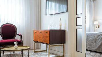 Habitación de hotel con un sillón rojo, un mueble de madera con dos cajones, una mesita redonda con un florero, un televisor montado en la pared y una cama con una colcha estampada.