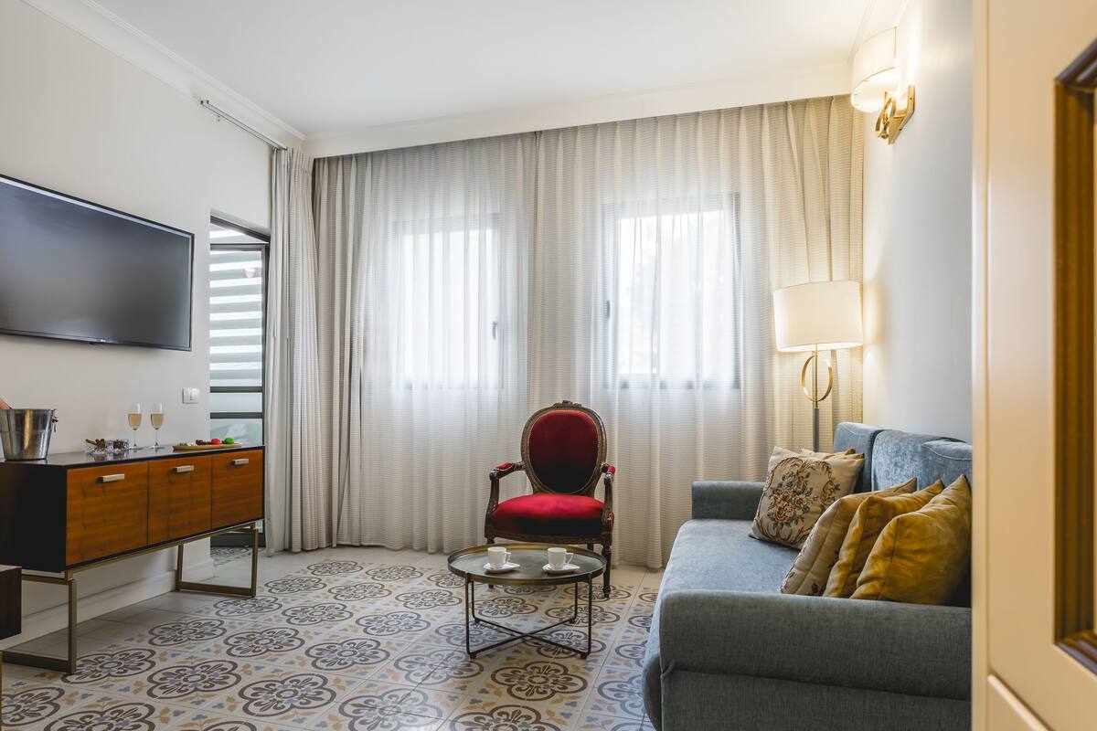 Un salon comprenant un canapé, un fauteuil rouge, une petite table basse ronde, un téléviseur fixé au mur et une lampe.