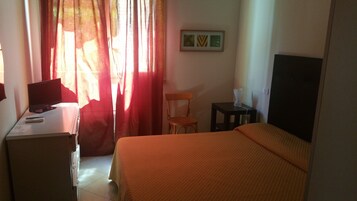 A hotel room with a bed, a desk with a computer, a chair, and a small table with a lamp.