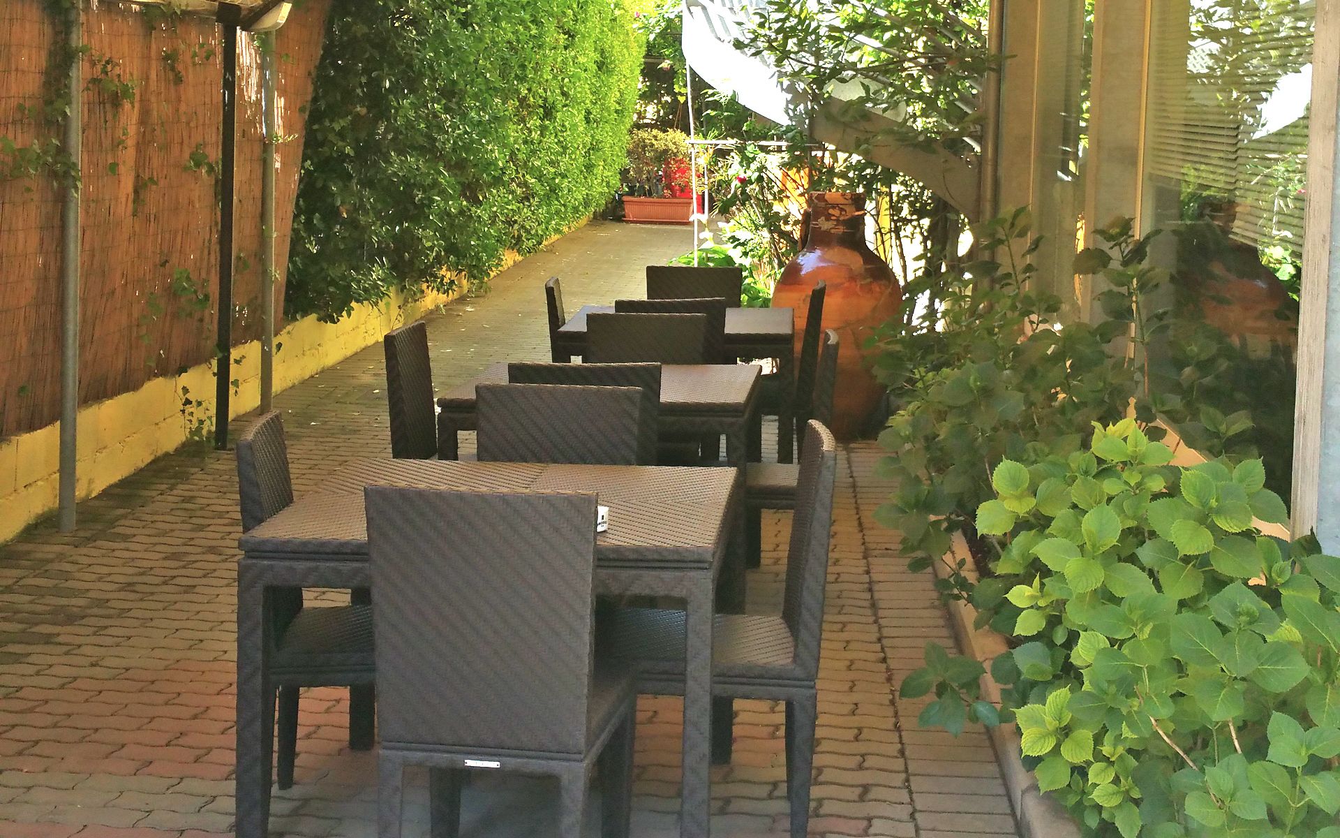 Un patio al aire libre con mesas y sillas de mimbre, rodeado de follaje y un camino de ladrillo.