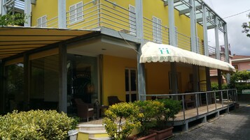 A yellow building with a covered outdoor seating area, a sign reading "HOTEL", and a small garden with potted plants.