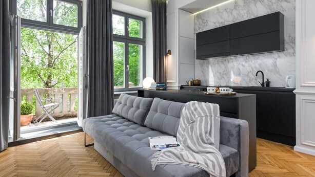 Ein modernes Wohnzimmer mit einer grauen Couch, einem Essbereich mit einer schwarzen Kücheninsel und einem Balkon mit Blick auf Bäume.