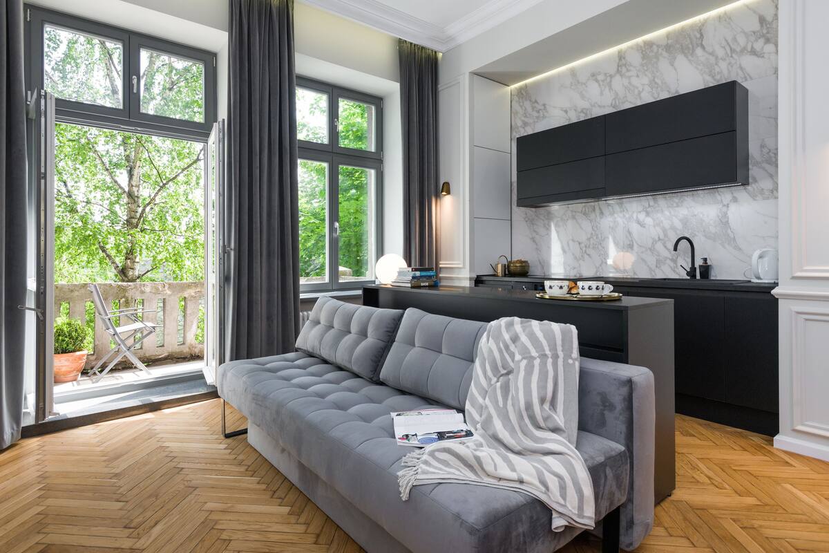Un salon moderne avec un canapé gris, un coin repas doté d’un îlot de cuisine noir, et un balcon offrant une vue sur les arbres.