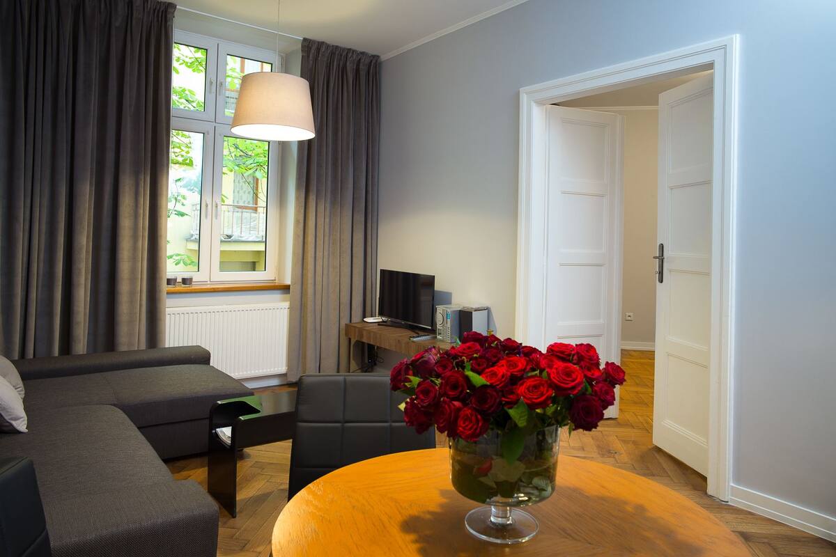 Un salon moderne avec un canapé, une table basse ornée d’un vase de roses rouges, un téléviseur et une fenêtre avec des rideaux.