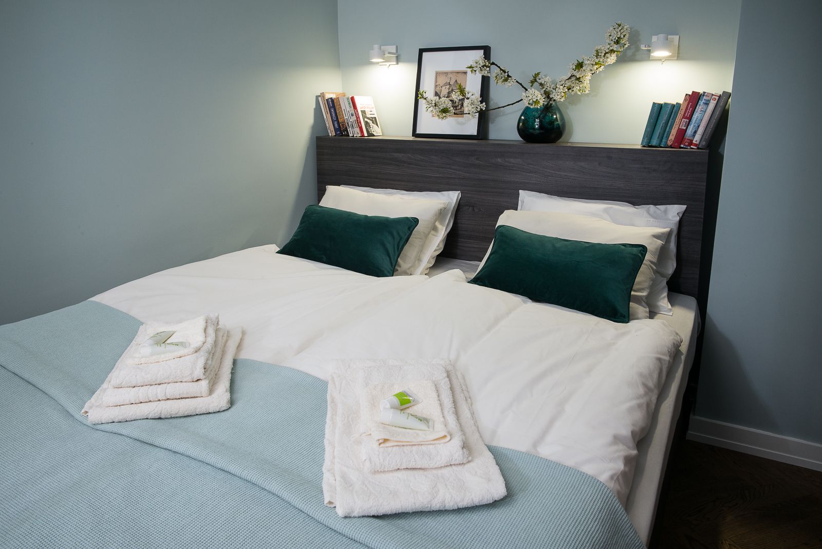 A neatly made bed with white linens and two green pillows, a vase with flowers, and a bookshelf with books.