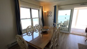 A dining area with a wooden table and white chairs, a bowl of fruit, and a view of the ocean through large windows. - Calypso unit 701 located at 62nd street and Ocean front (Ocean City)