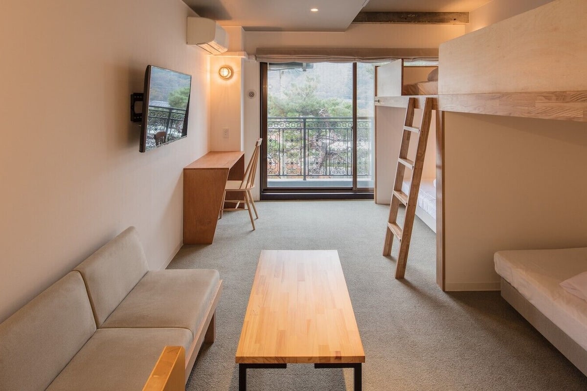 A room with a sofa, a wooden table, a bunk bed, a ladder, a desk, and a balcony with a railing.