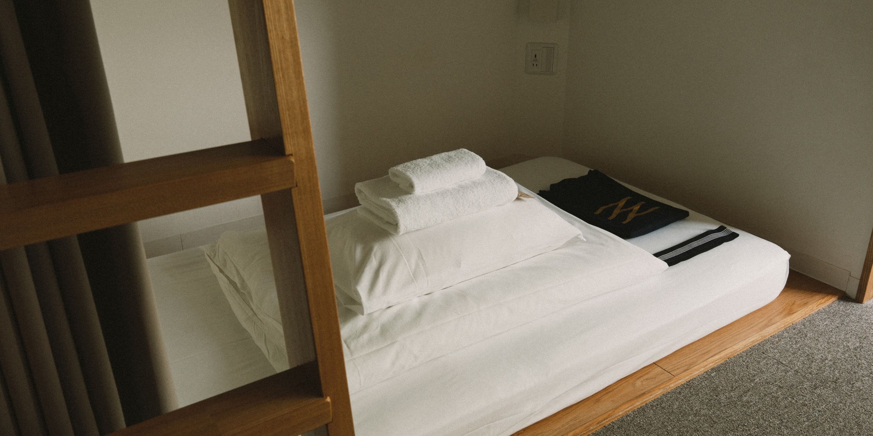 Un lit simple avec une literie blanche et un oreiller noir orné d’un écusson doré, un plancher en bois et un mur blanc avec une prise de courant.