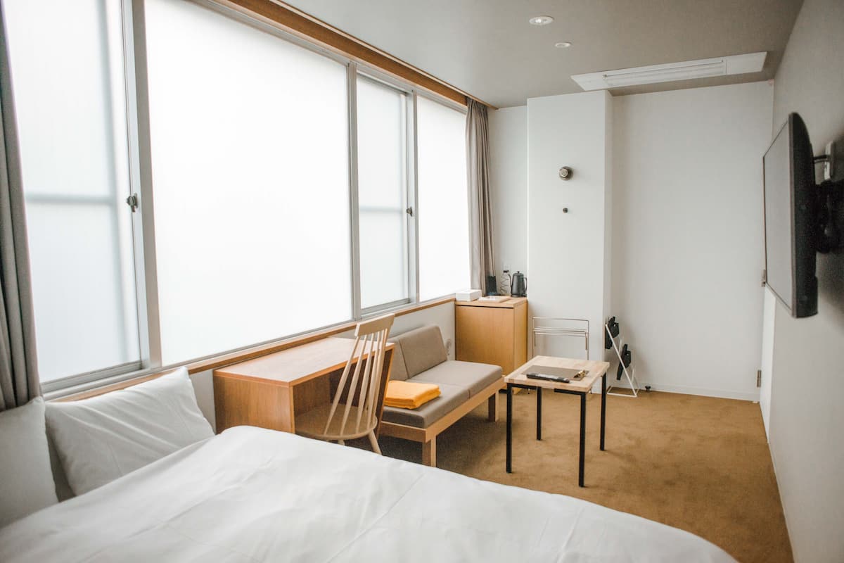 A hotel room with a bed, a desk, a chair, a small table, and a TV mounted on the wall.