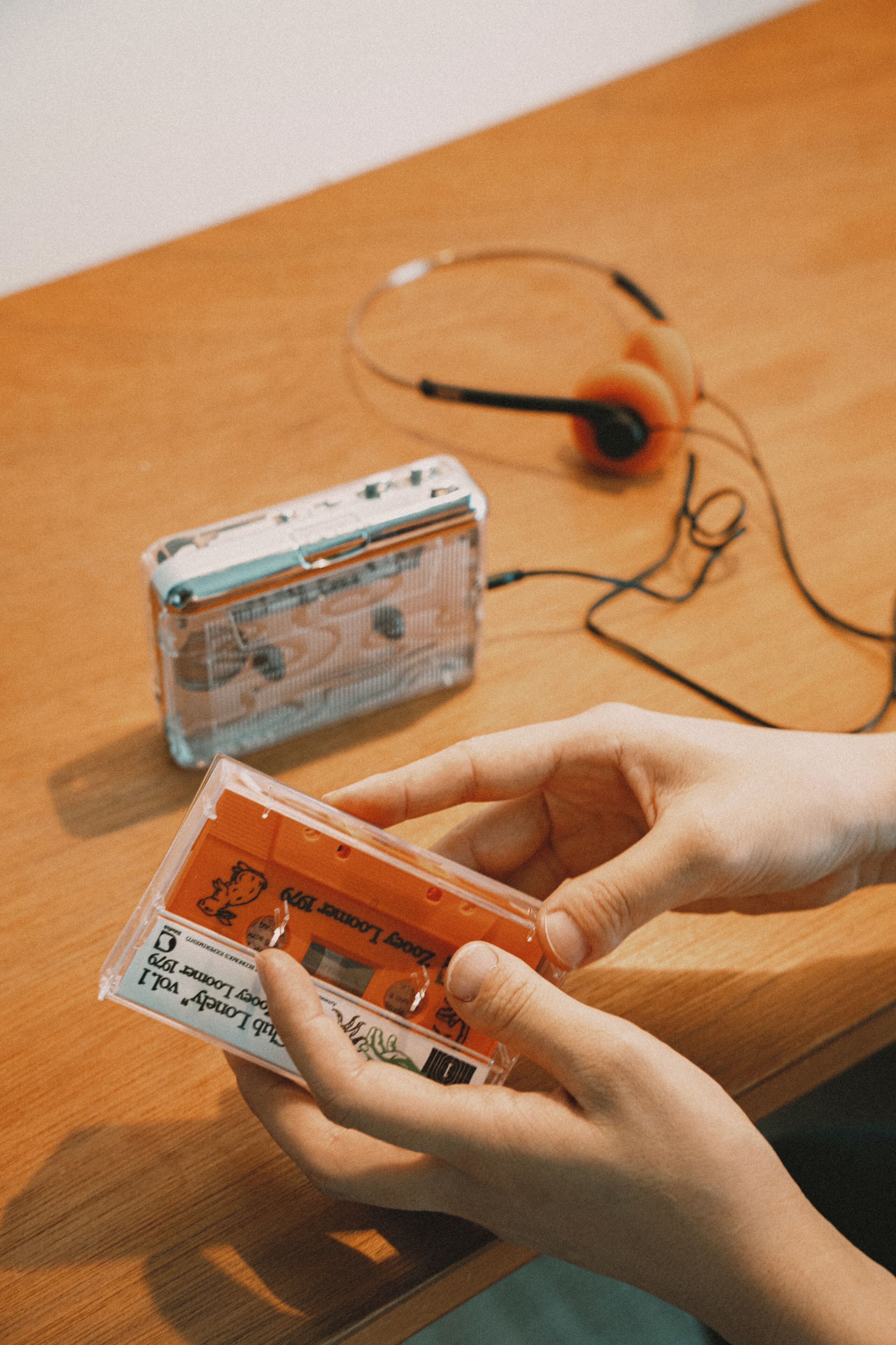 A person holding a cassette tape with a label, a transparent case, and a pair of headphones on a wooden surface.