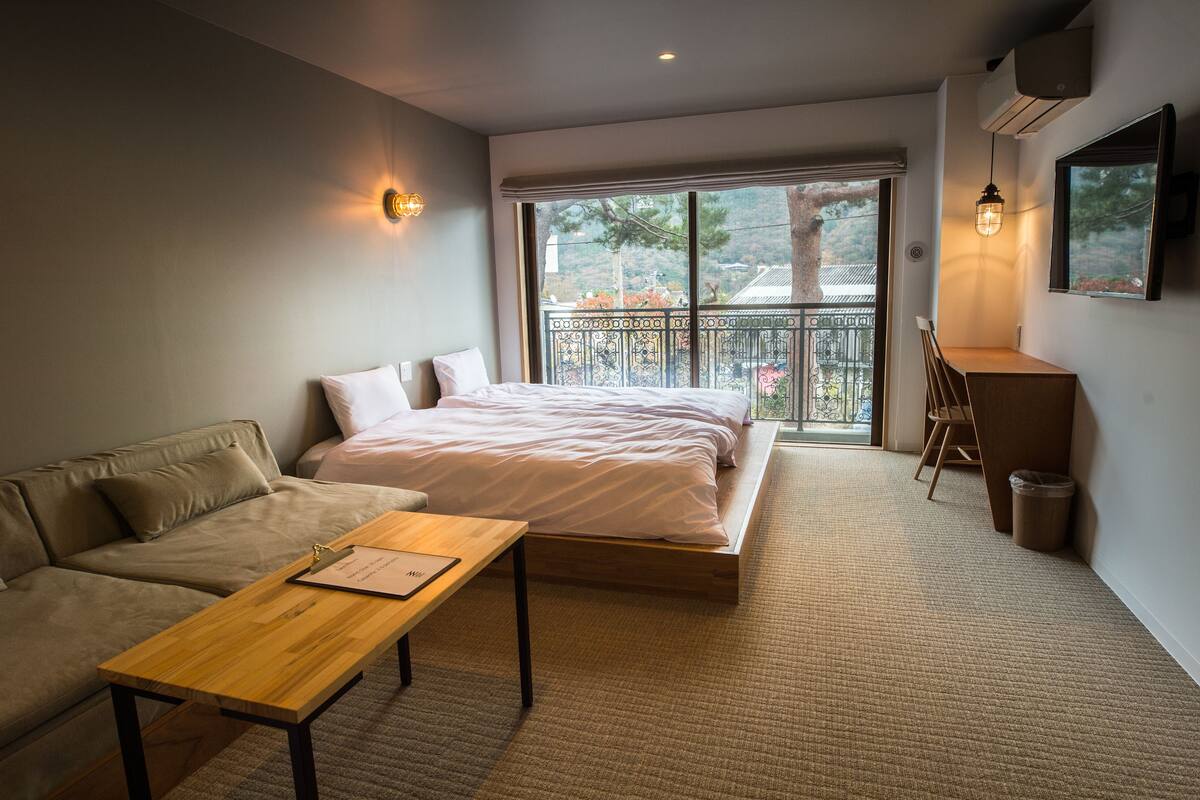 A modern bedroom with a bed, a sofa, a desk, and a balcony with a view of trees.