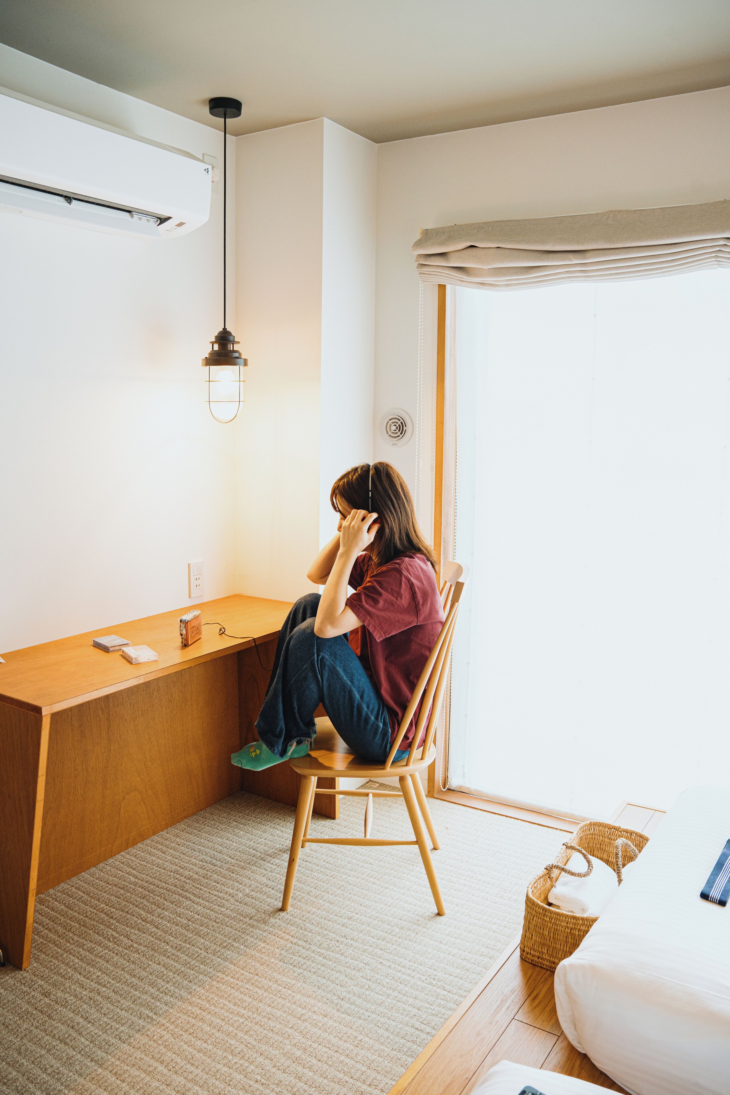 A room with a wooden desk, a chair, a bed, a window with curtains, and a hanging light fixture.