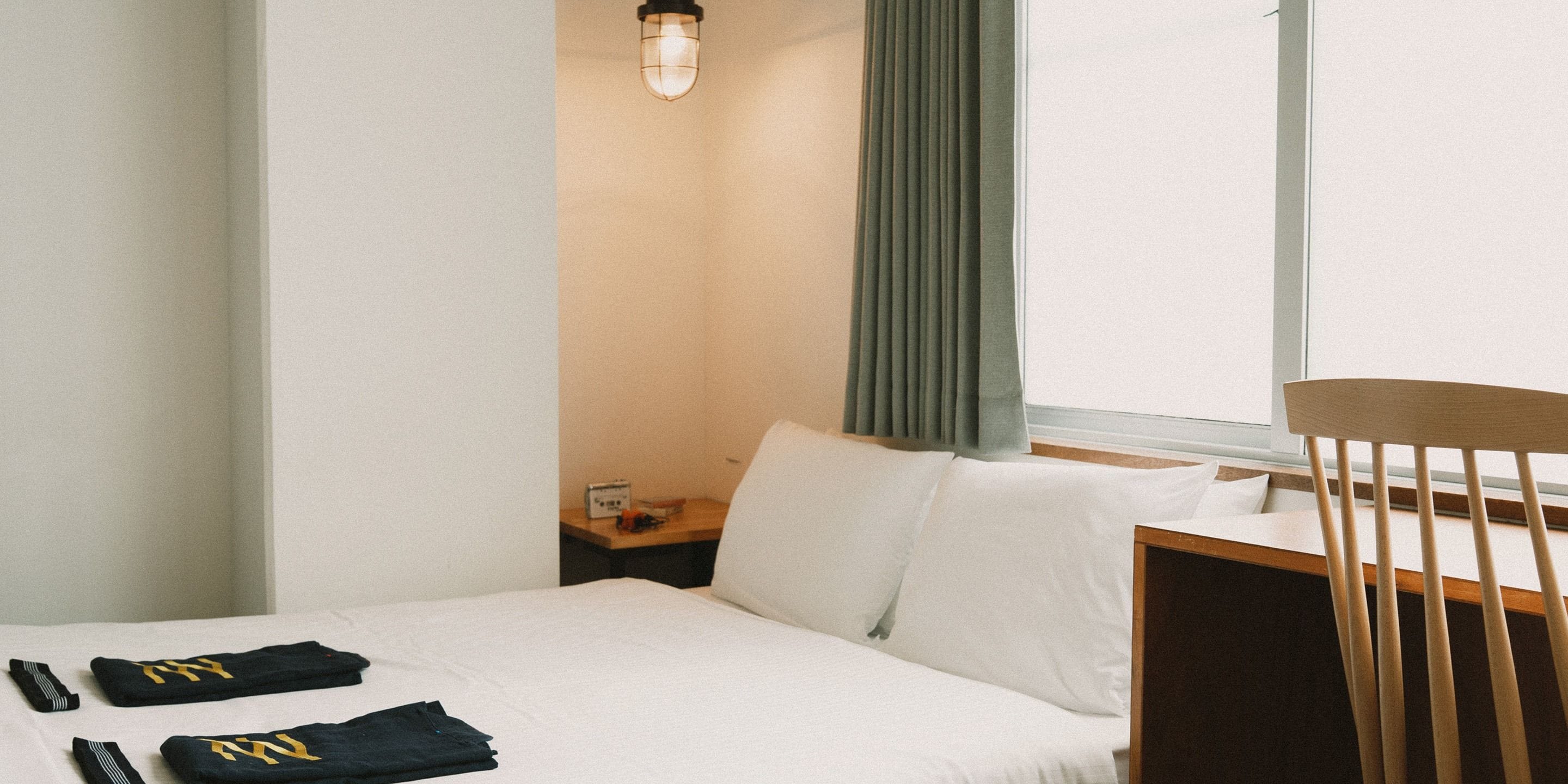 A hotel room with a bed, a wooden chair, a nightstand with a lamp, and a window with curtains.