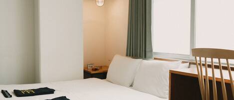 A hotel room with a bed, a wooden chair, a nightstand with a lamp, and a window with curtains.