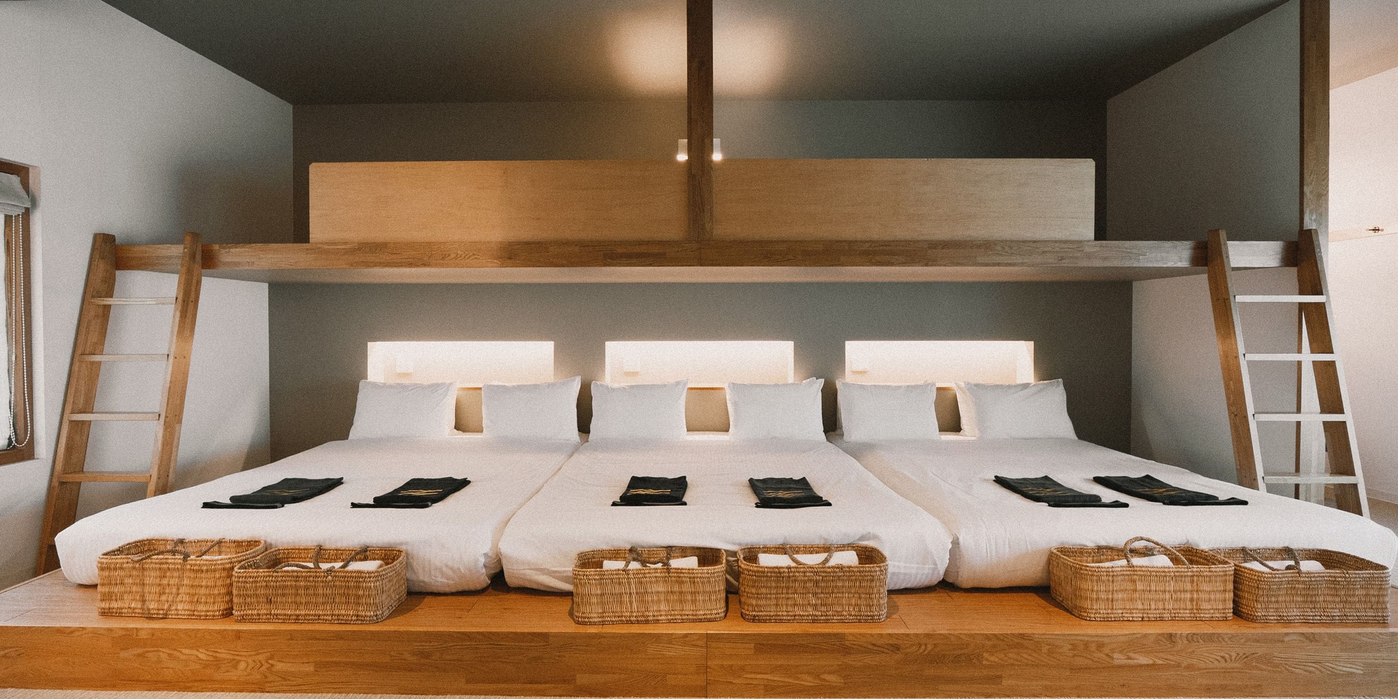 A room with three beds arranged in a row, each with a headboard, pillows, and a blanket. There are wooden ladders on either side of the beds, and baskets placed at the foot of each bed.