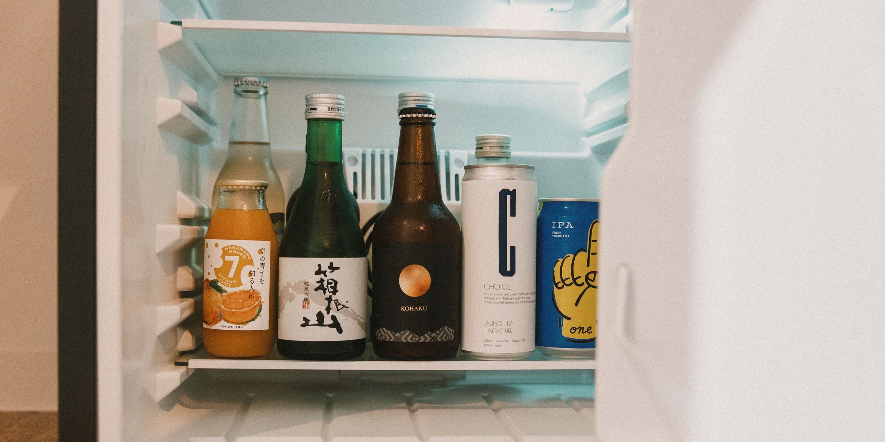 A refrigerator shelf with various beverages including a bottle of citrus juice, a bottle of hard cider, a can of beer, and a can of soft drink.