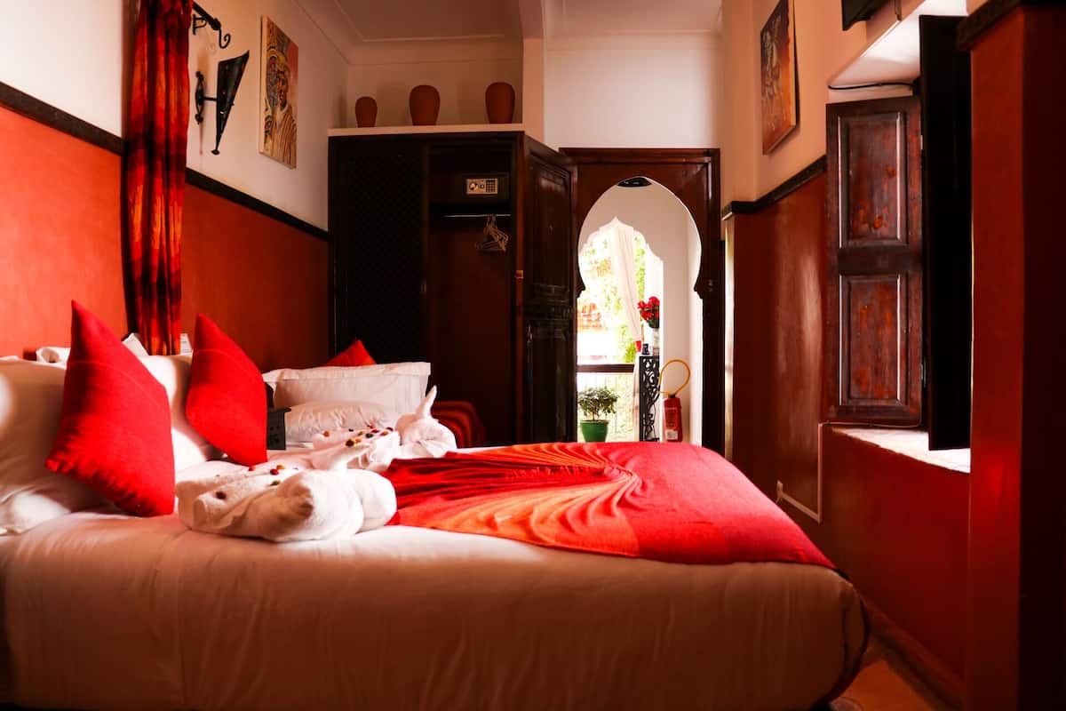 A hotel room with a large bed, red pillows, a red blanket, and a view of a courtyard through an open door.