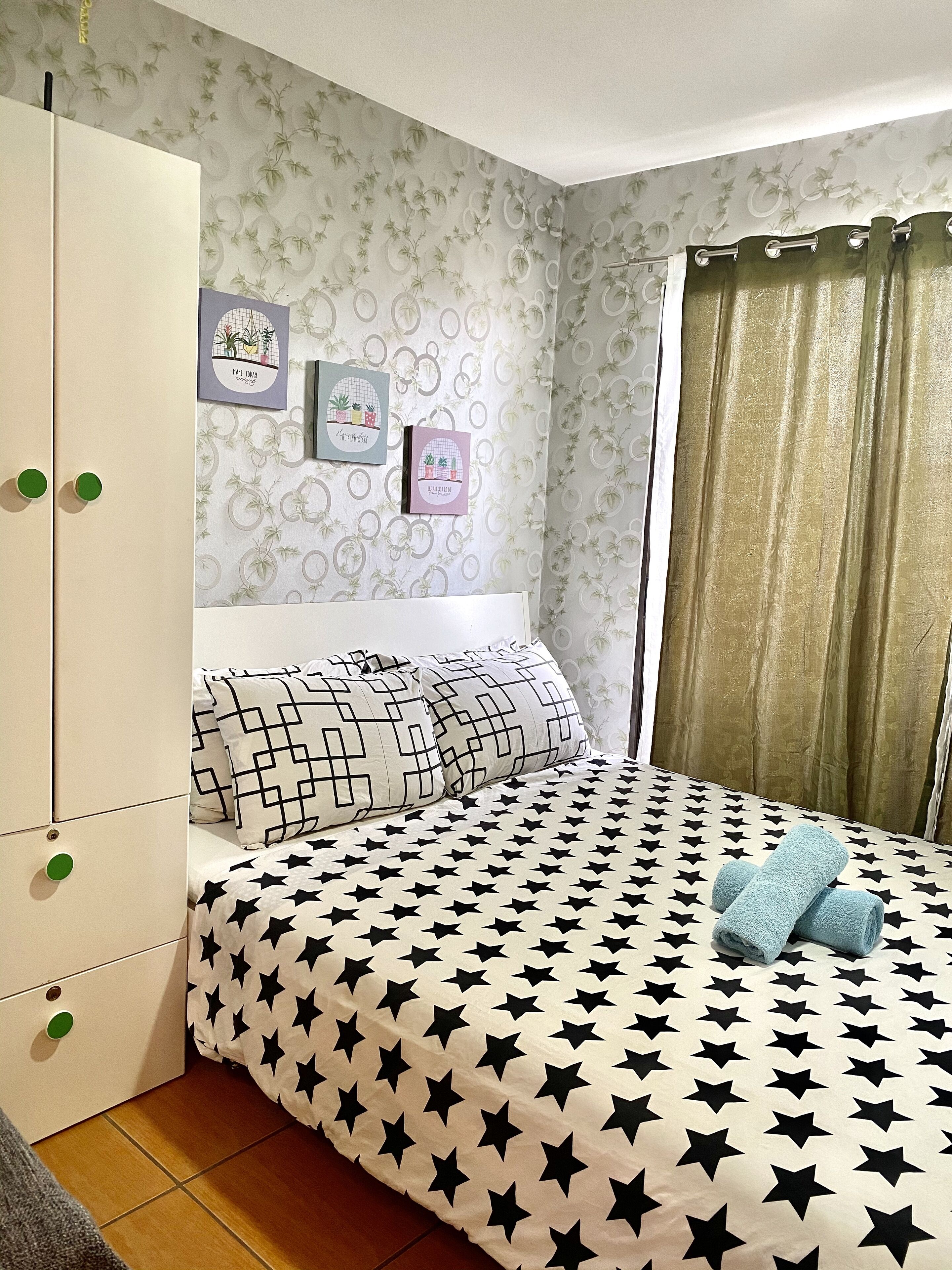 A bedroom with a bed featuring a black and white star pattern, a wardrobe with green handles, and a wall with framed botanical prints.