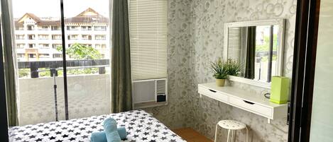 A hotel room with a bed, a bedside table with a mirror, a potted plant, and a view of a building outside.