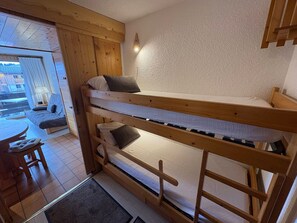 Un lit superposé avec une structure en bois, une petite table et une chaise, dans une pièce dont le sol est carrelé et qui offre une vue à travers une porte vitrée.