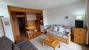 Un espace de vie compact comprenant un canapé, une table basse en bois, un canapé à motifs, un téléviseur et un coin cuisine.