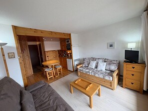 Een knus woongedeelte met een bank, een houten bijzettafeltje, een gestreept bankje, een televisie en een kitchenette.