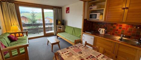 Et hyggeligt værelse med en grøn sofa, et træbord og et tekøkken med mikroovn og vask.