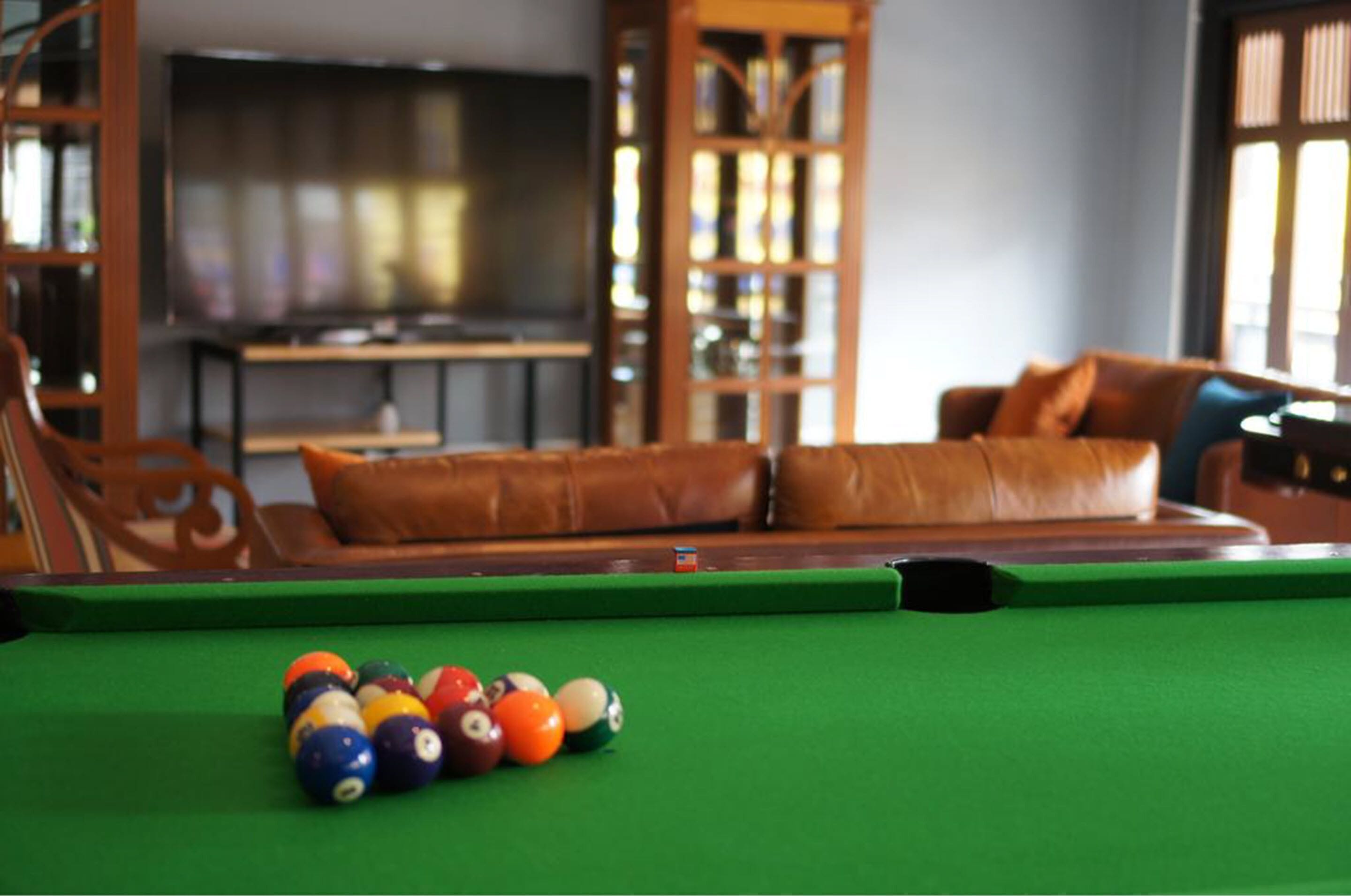 A pool table with balls in the foreground, a brown leather sofa, and a wooden cabinet with glass doors in the background.