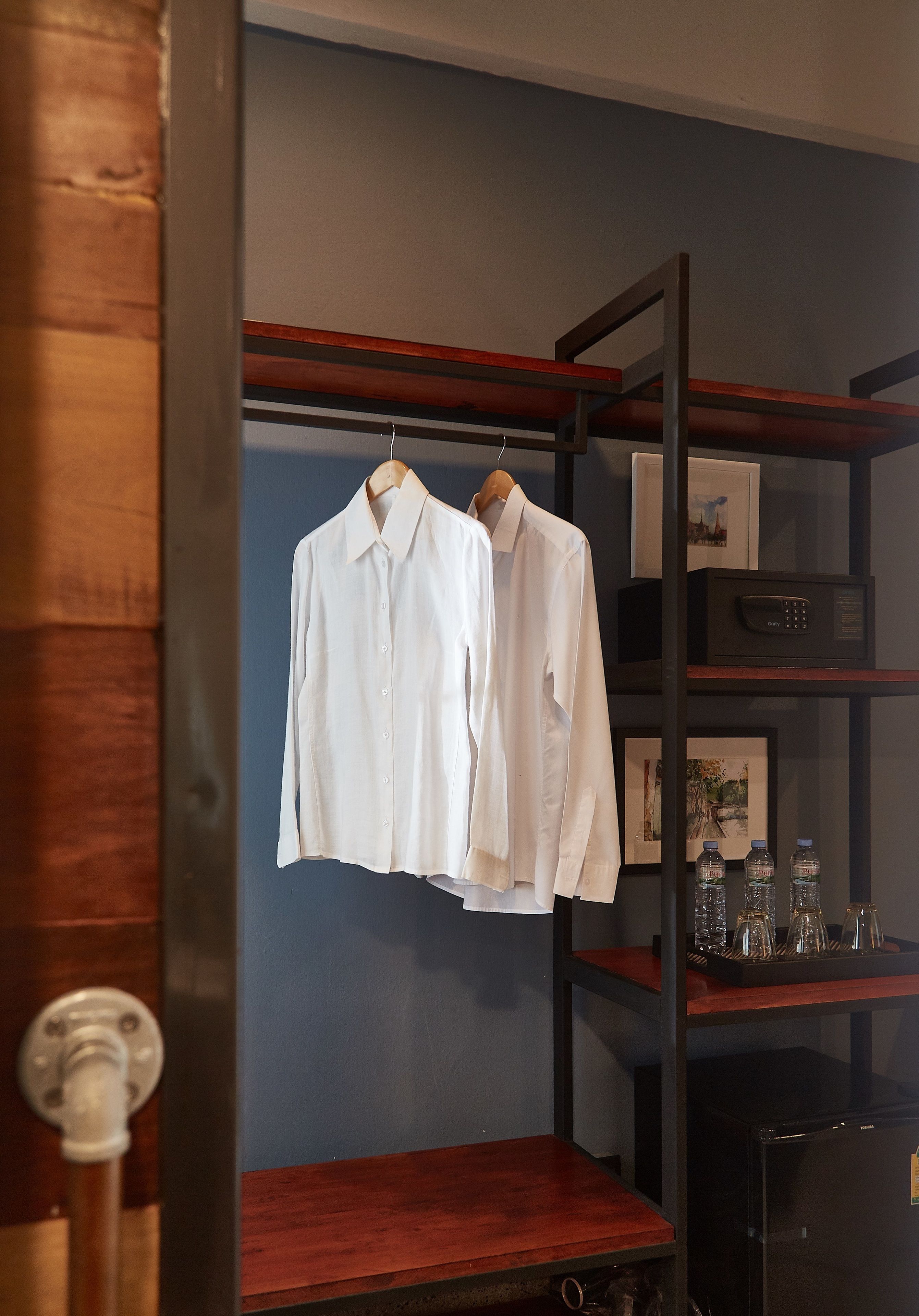 Une pièce avec une armoire en bois, un portant sur lequel sont suspendues deux chemises blanches, et une étagère avec des bouteilles et des verres.
