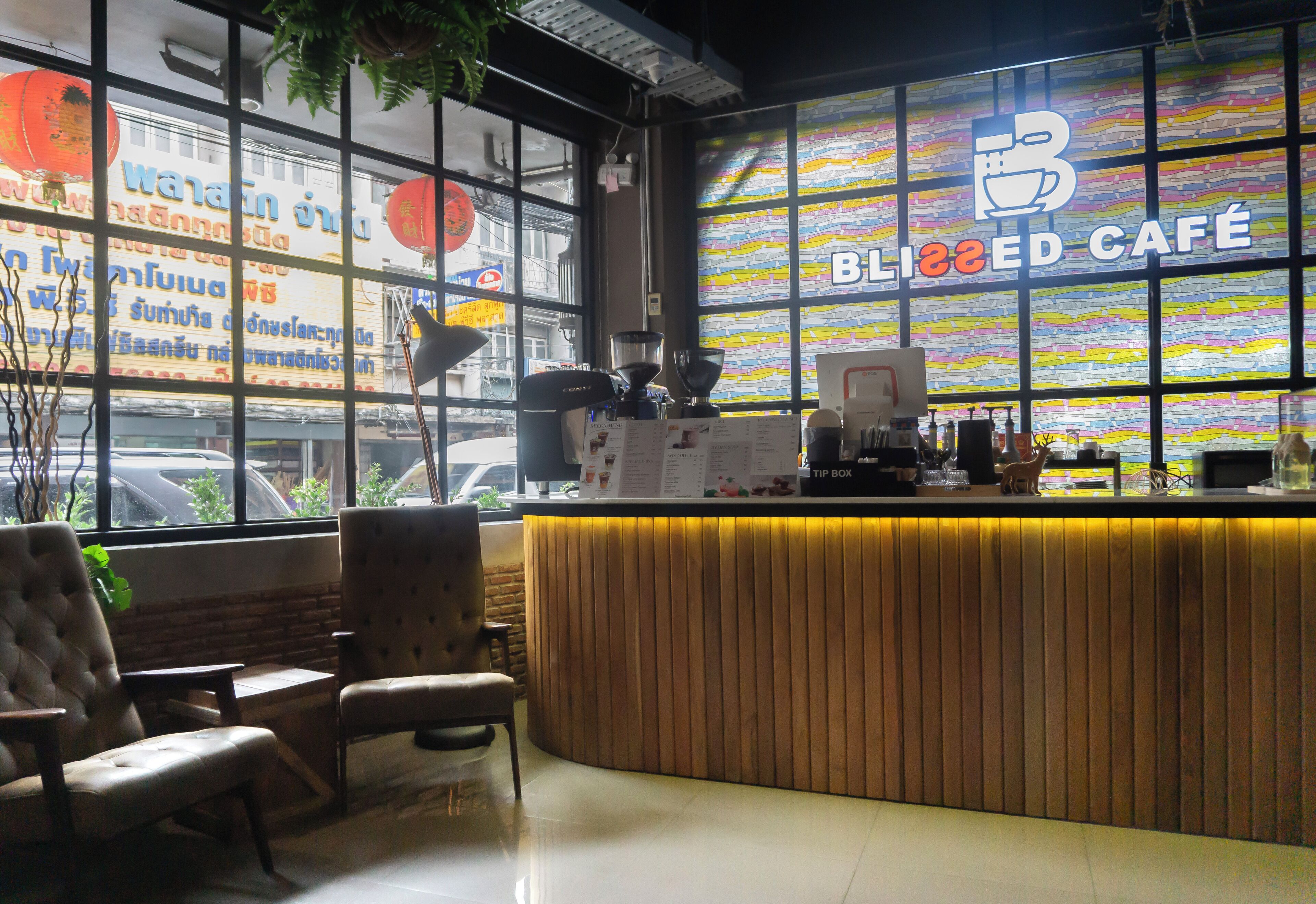 Un café doté d’un comptoir en bois, d’un espace salon et de grandes fenêtres offrant une vue colorée sur l’extérieur.