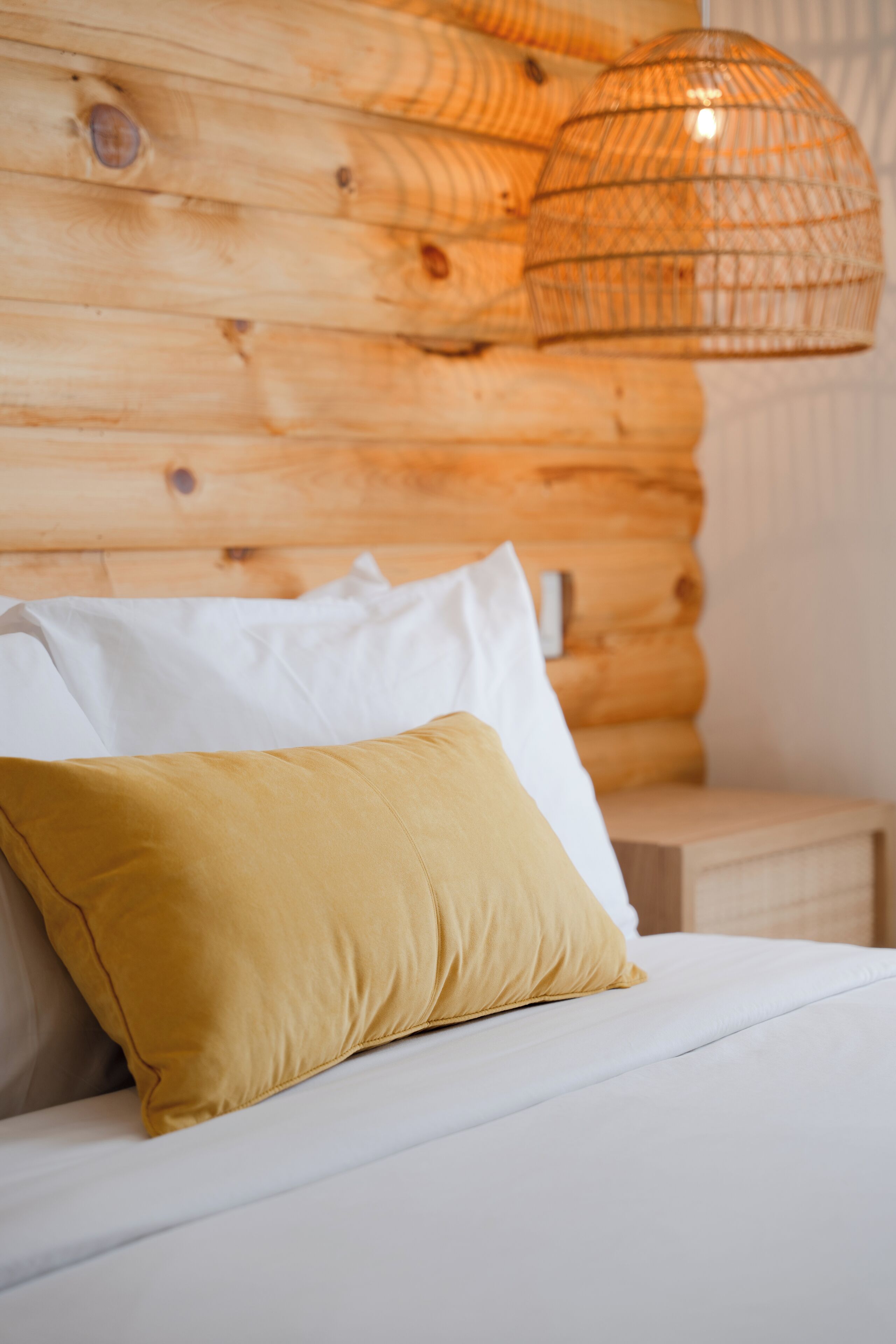 A bed with white and mustard pillows, a wooden headboard, and a woven light fixture.