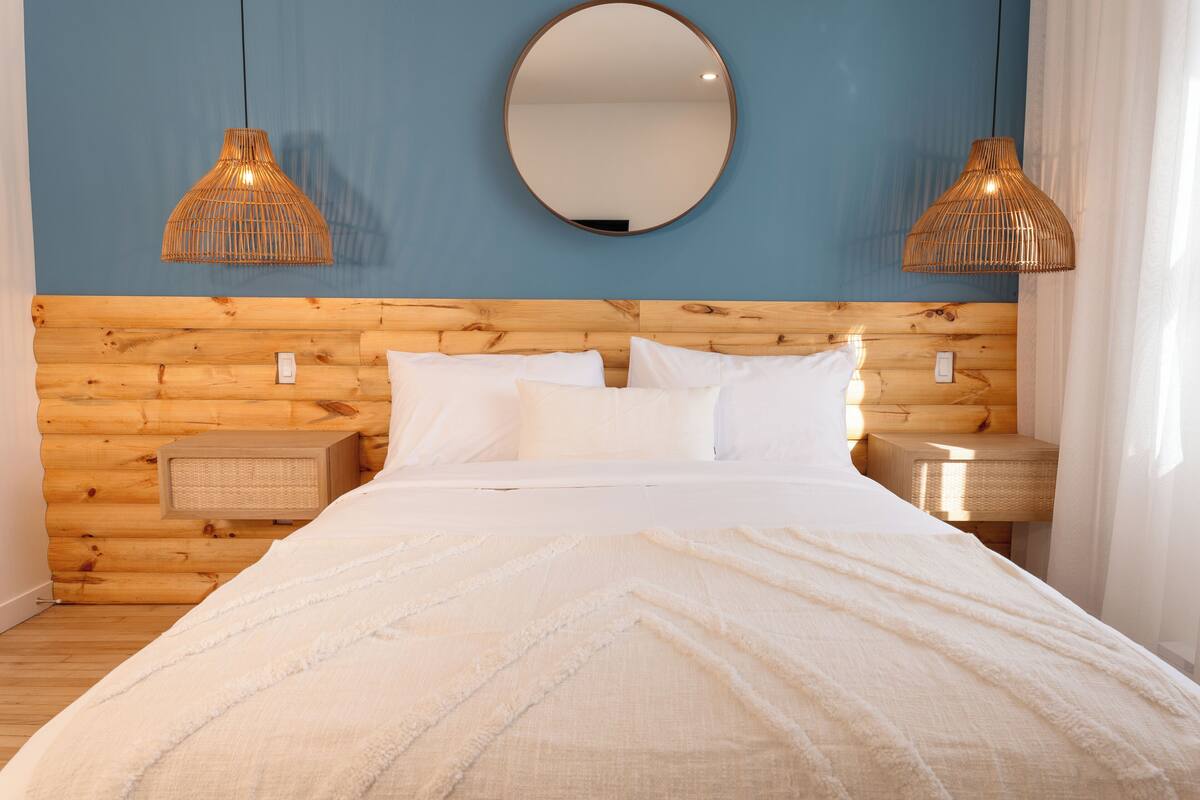 Un dormitorio con cabecera de madera, un espejo redondo, dos lámparas de noche y una cama con ropa de cama blanca.
