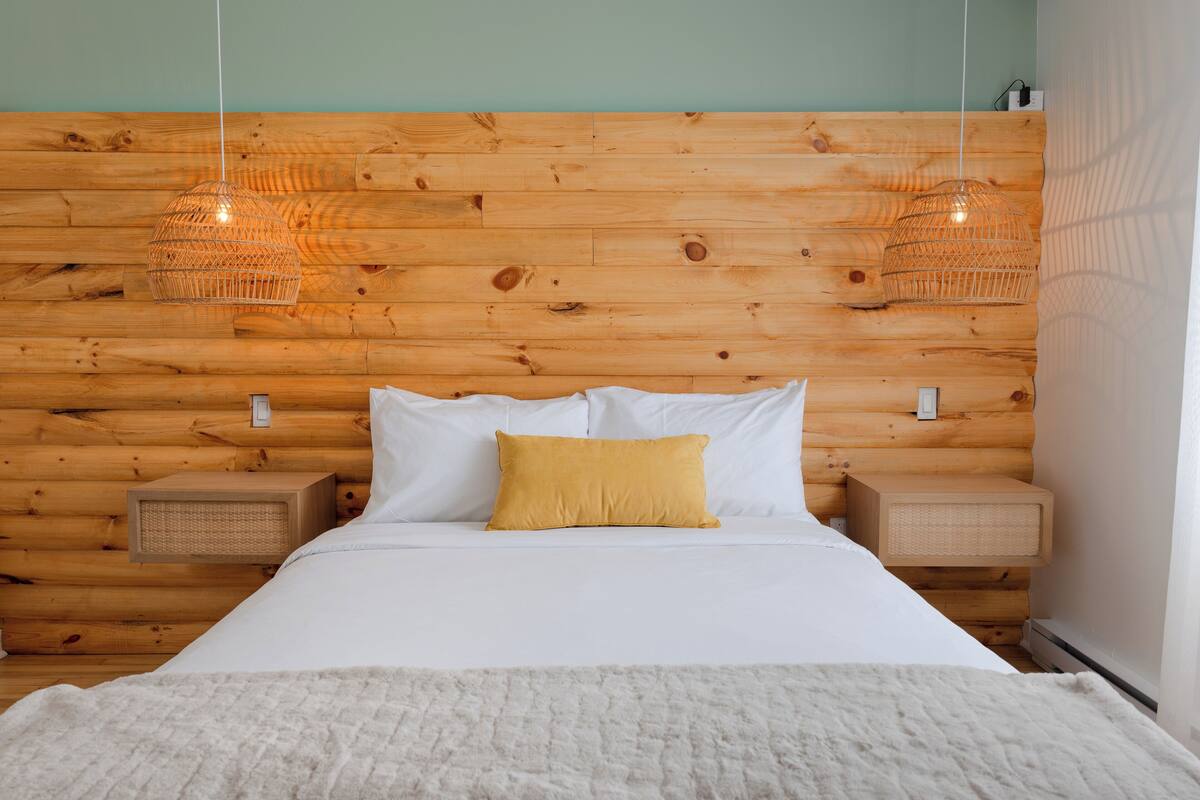 Un dormitorio con cabecera de madera, ropa de cama blanca, almohada amarilla y dos mesitas de noche con cestas tejidas.