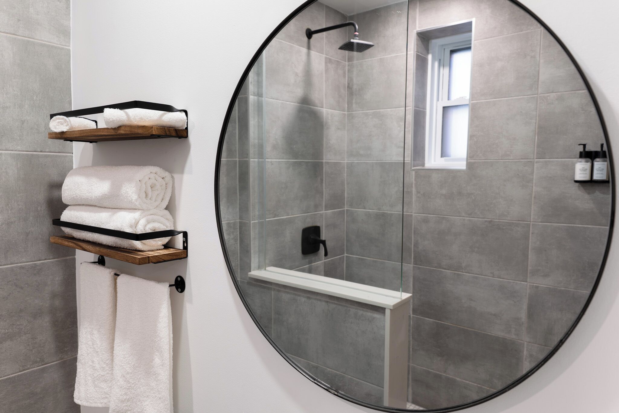 Un baño moderno con baldosas grises, una ducha con mampara de vidrio, una bañera blanca y un perchero con toallas dobladas con cuidado.