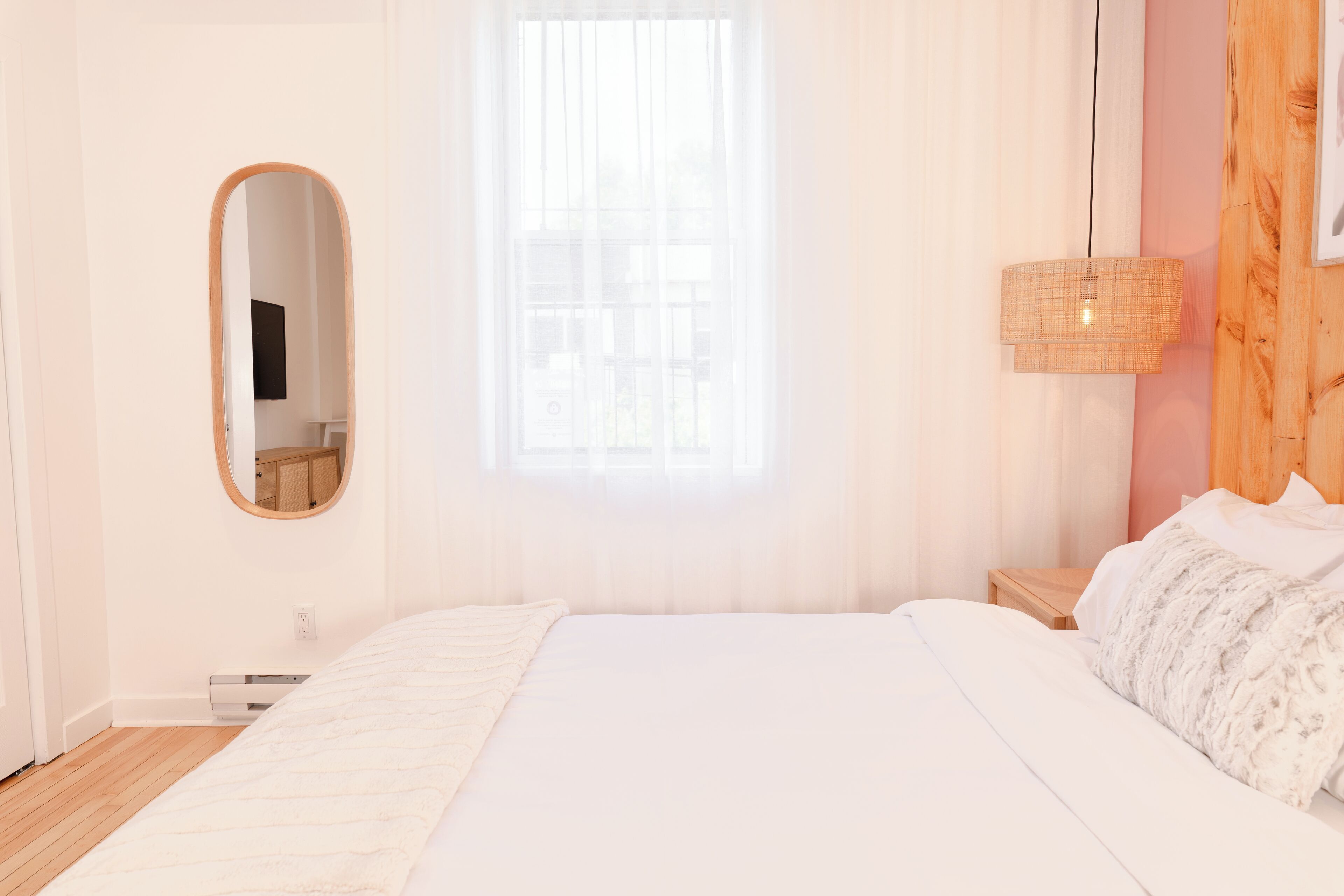 Un dormitorio con una cama grande, una mesita de noche, un espejo, una ventana con cortinas translúcidas y una lámpara de pared.
