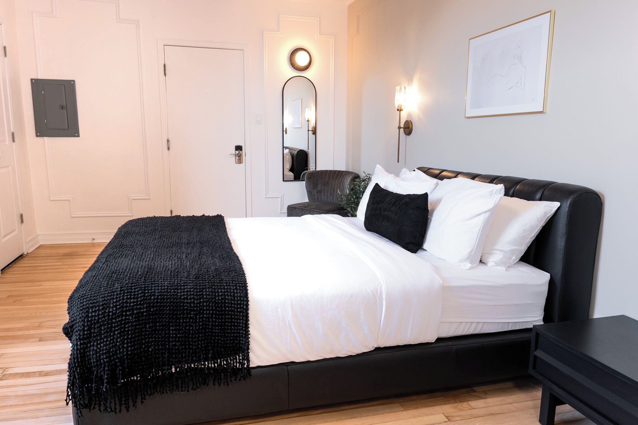 A modern bedroom with a black bed, white bedding, a black throw blanket, a nightstand, a wall-mounted mirror, and a framed picture.