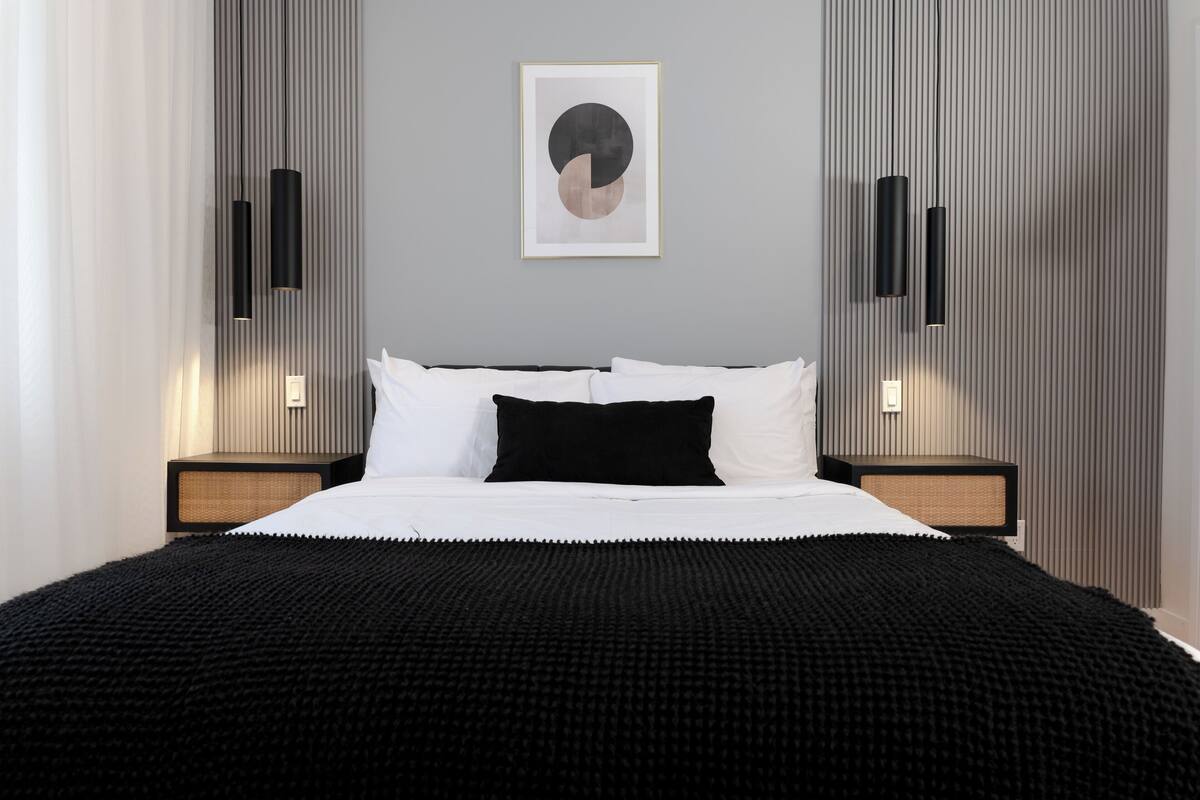 Una habitación de hotel moderna con una cama grande, dos mesitas de noche, una lámpara de pared y una obra de arte enmarcada sobre la cama.