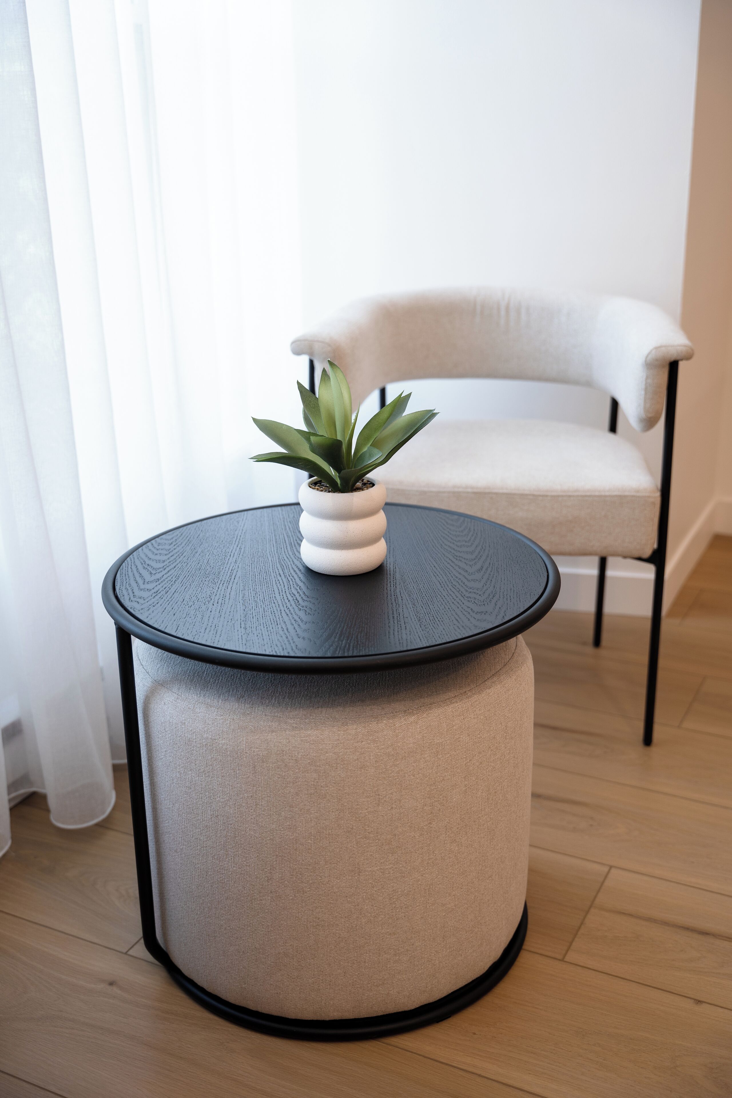 Une table ronde, un pot de fleurs, une chaise et une fenêtre ornée de rideaux légers.