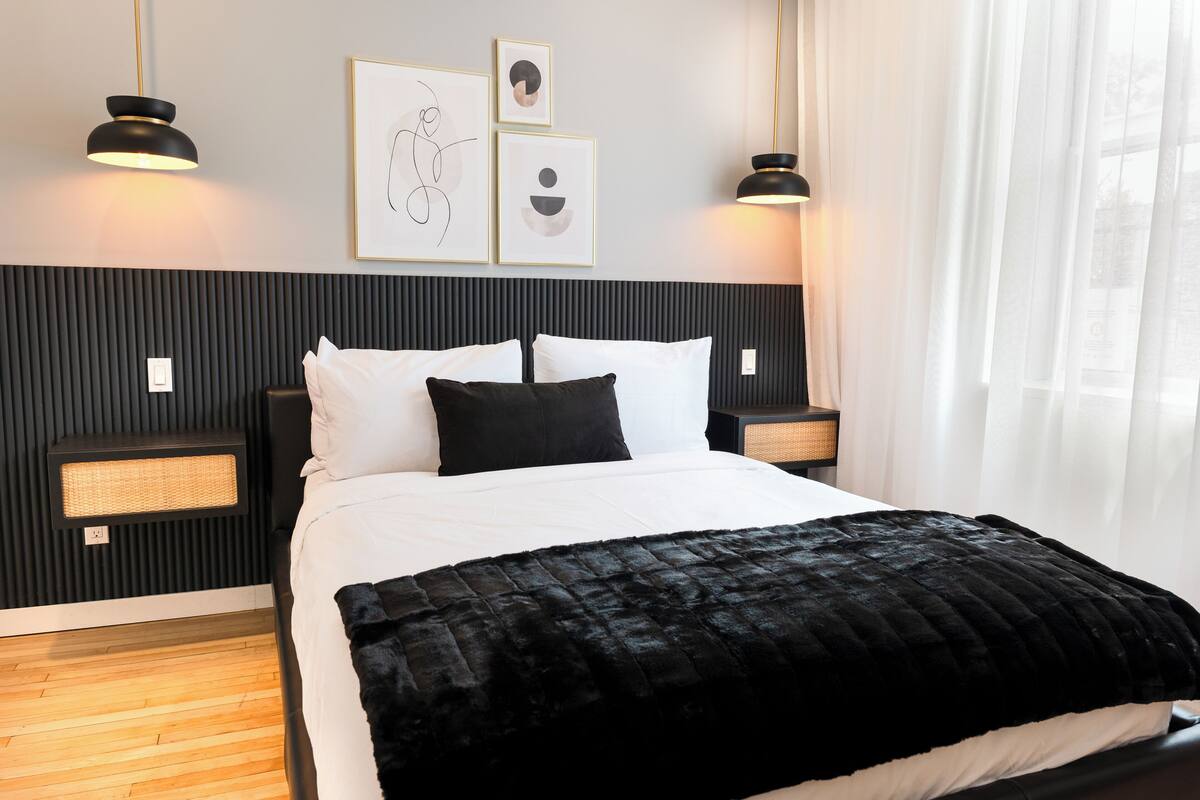 Un dormitorio moderno con cabecera negra, ropa de cama blanca y una manta negra. Hay dos mesitas de noche con lámparas, un calentador en la pared y cuadros enmarcados sobre la cama.