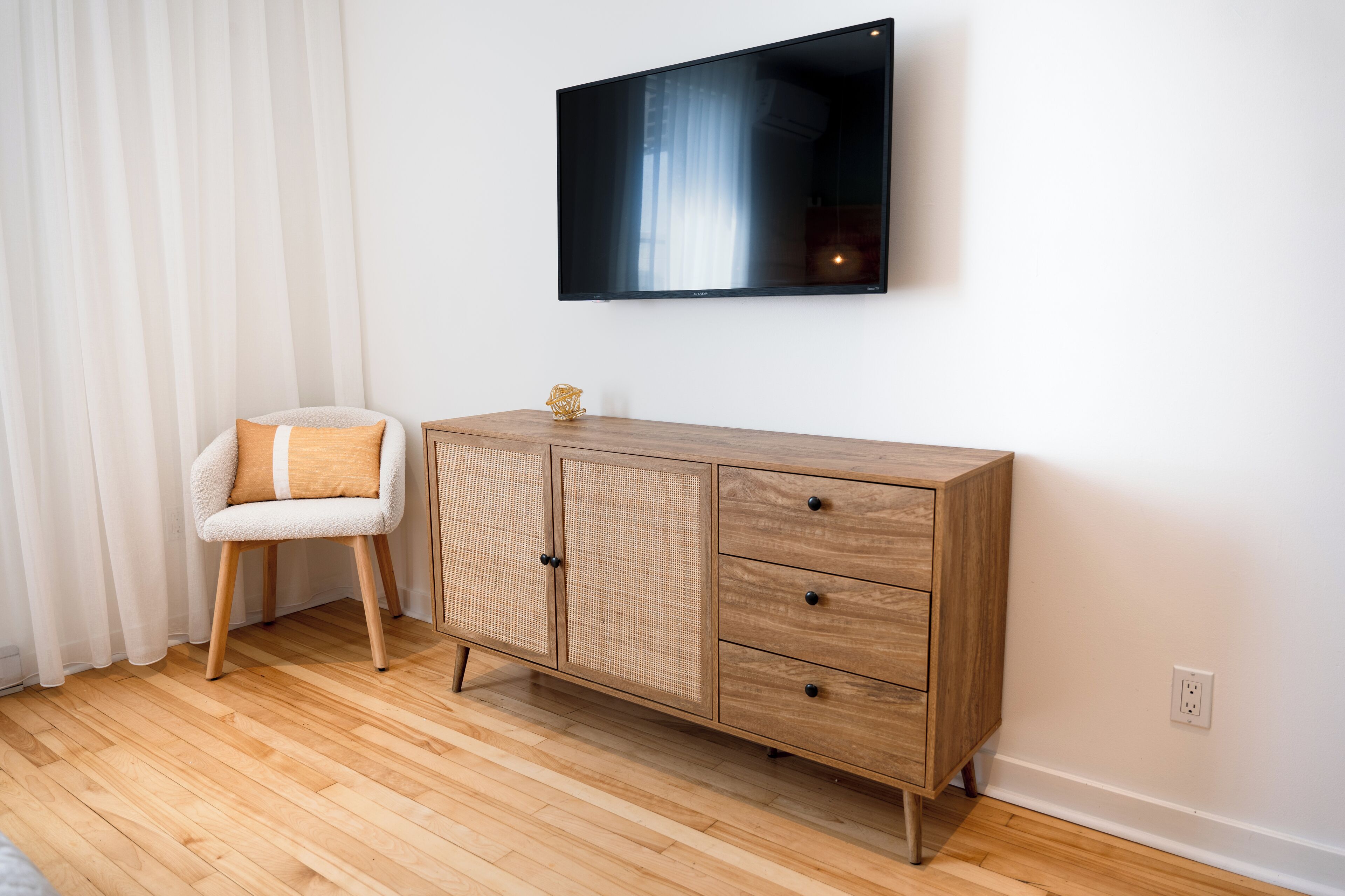 A room with a wooden cabinet, a white chair with a cushion, and a mounted television.