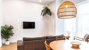 A modern living room with a grey sofa, wooden dining table, and chairs, a TV mounted on the wall, and a hanging plant.