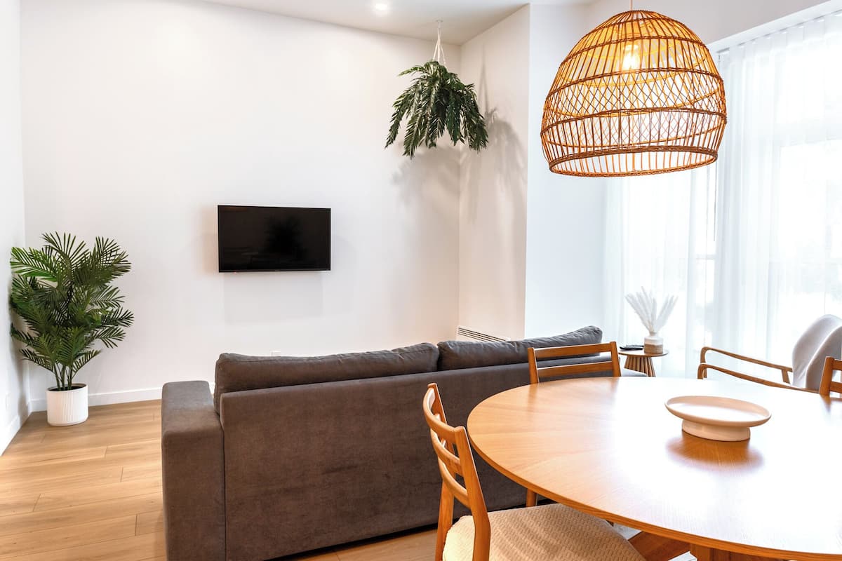 Una sala moderna con un sofá gris, una mesa de comedor de madera y sillas, un televisor montado en la pared y una planta colgante.
