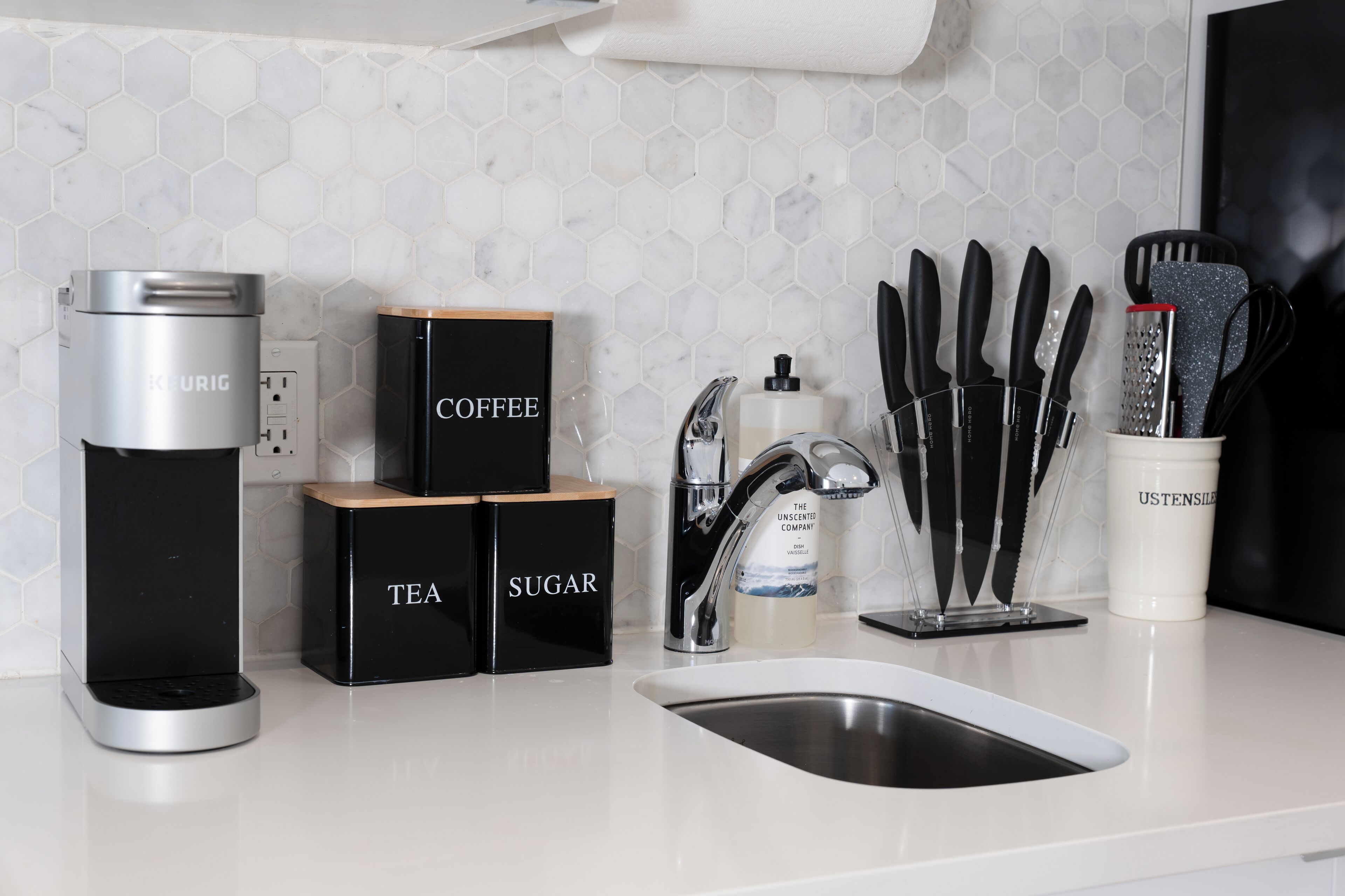 A kitchen with a coffee maker, tea and sugar containers, a sink, and utensils.