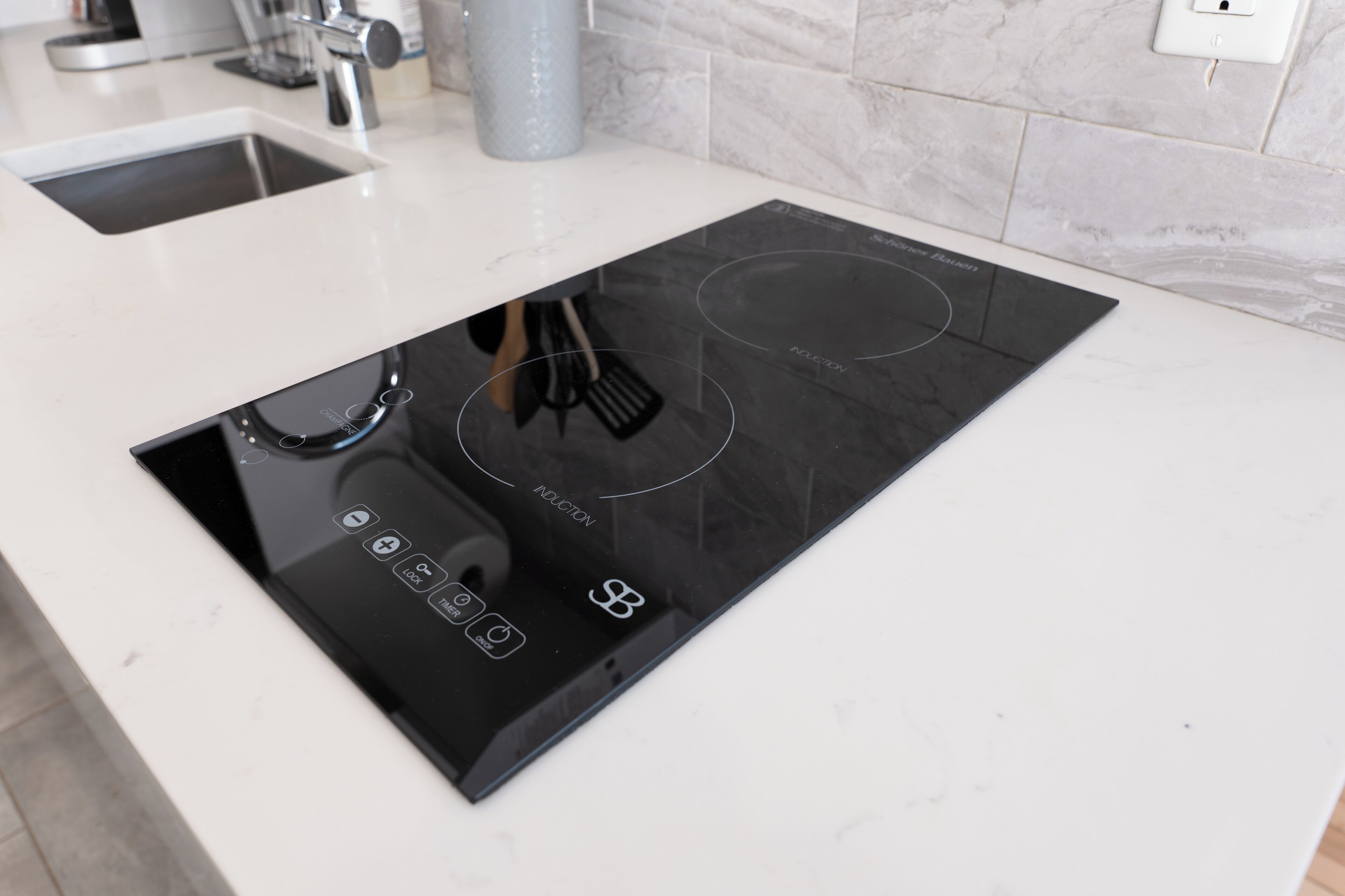 A modern kitchen with a black induction cooktop, a sink, and a marble backsplash.